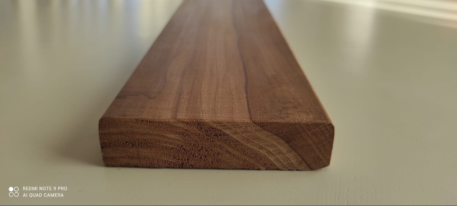 Wyprofilowana deska z ciemnego drewna o widocznych słojach, leżąca na jasnym tle. Widoczny przekrój i struktura drewna.