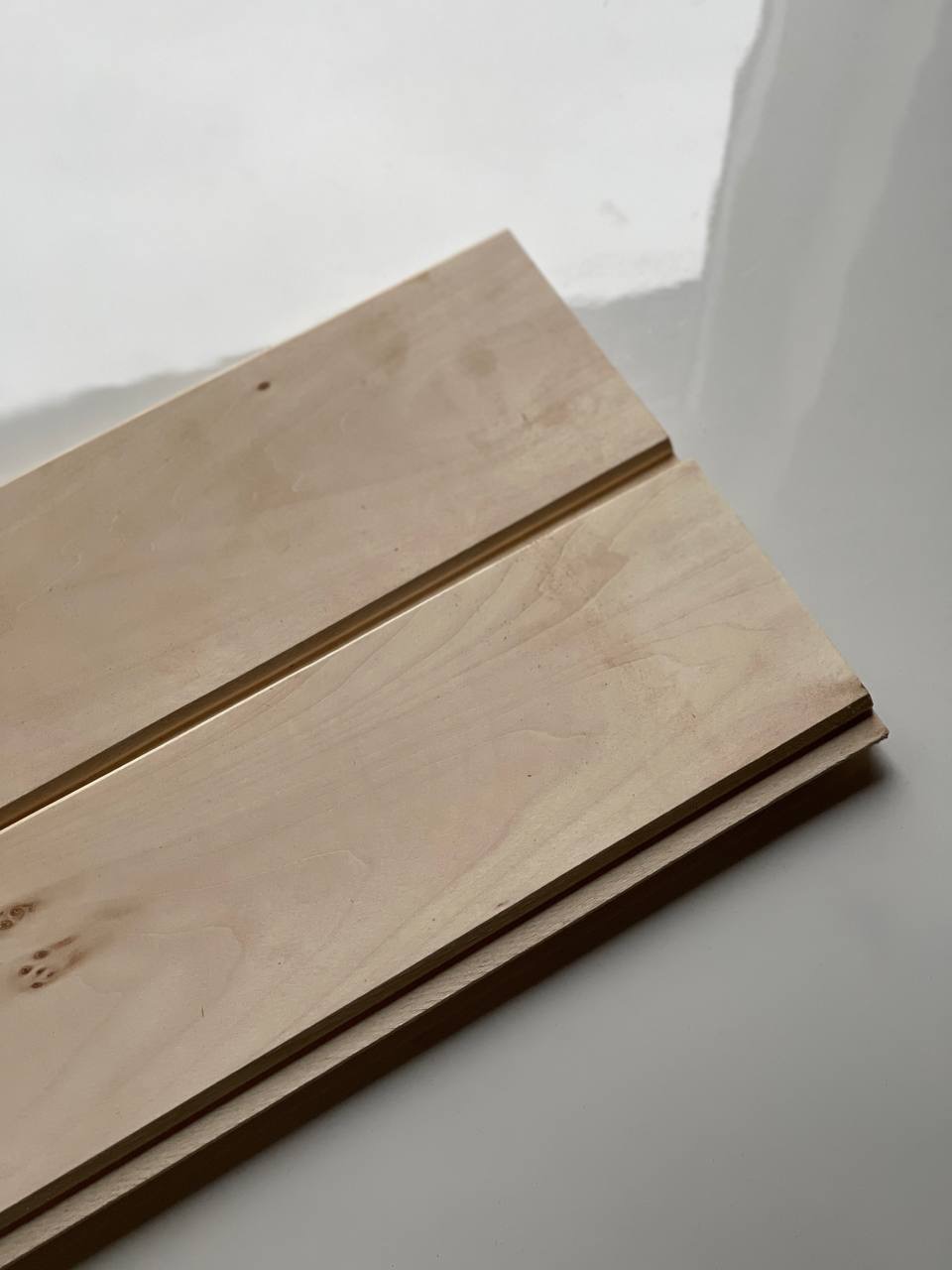 Dwie jasne, surowe deski z widocznym usłojeniem, ułożone jedna na drugiej. Widoczne krawędzie i delikatne sęki. Całość na jasnym, błyszczącym tle.