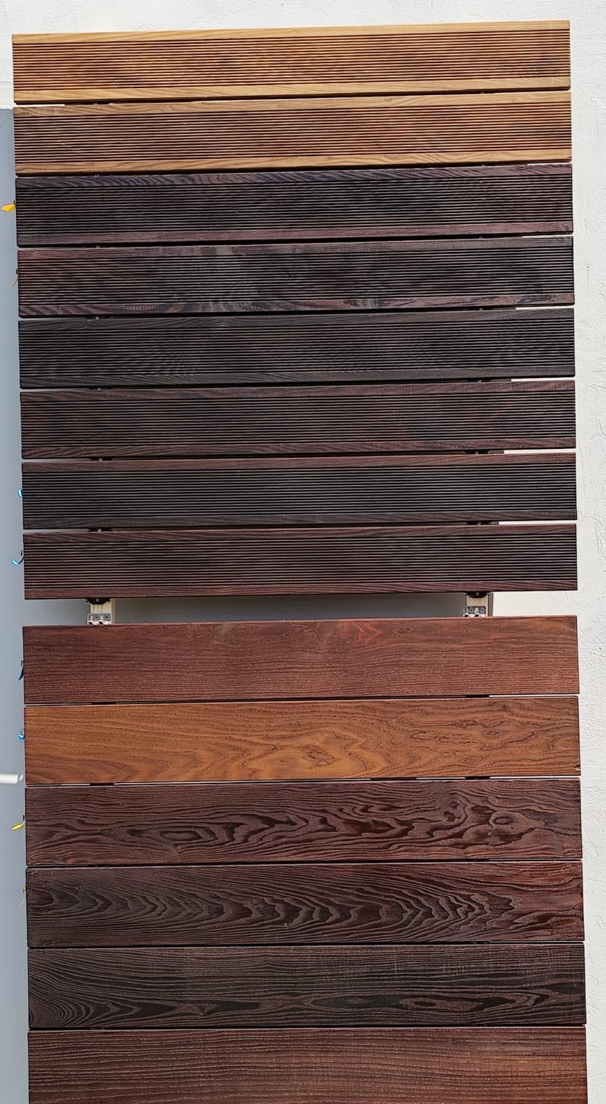 Ekspozycja różnych odcieni desek tarasowych z drewna egzotycznego, zamontowanych na metalowych wspornikach na jasnym tle. Widoczne zróżnicowane faktury i kolory.