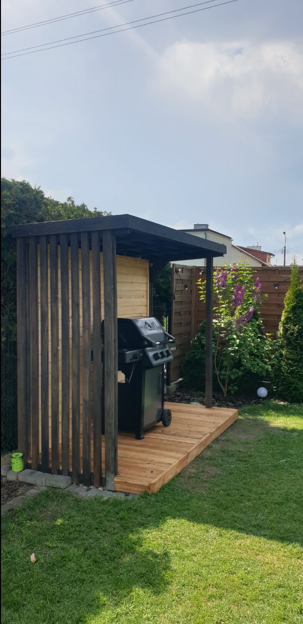 Zadaszona, drewniana konstrukcja z grillem na tarasie z desek. Widoczne pionowe deski, roślinność i trawnik. Całość utrzymana w jasnej kolorystyce z ciemnymi akcentami.