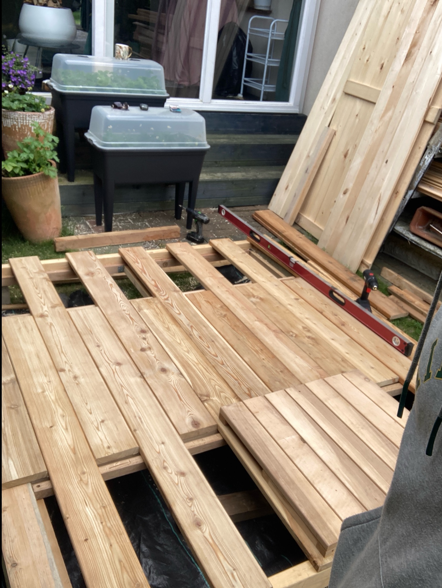 Budowa drewnianego tarasu: deski ułożone na legarach, poziomica i wkrętarka w tle. Częściowo ukończona konstrukcja z widoczną czarną folią pod spodem.