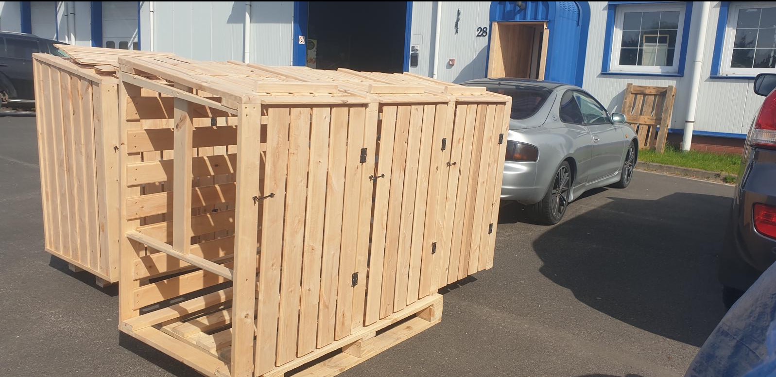 Dwie drewniane konstrukcje z zamykanymi drzwiczkami, prawdopodobnie na śmietniki, stoją na paletach przed budynkiem z niebieskimi drzwiami i srebrnym samochodem.