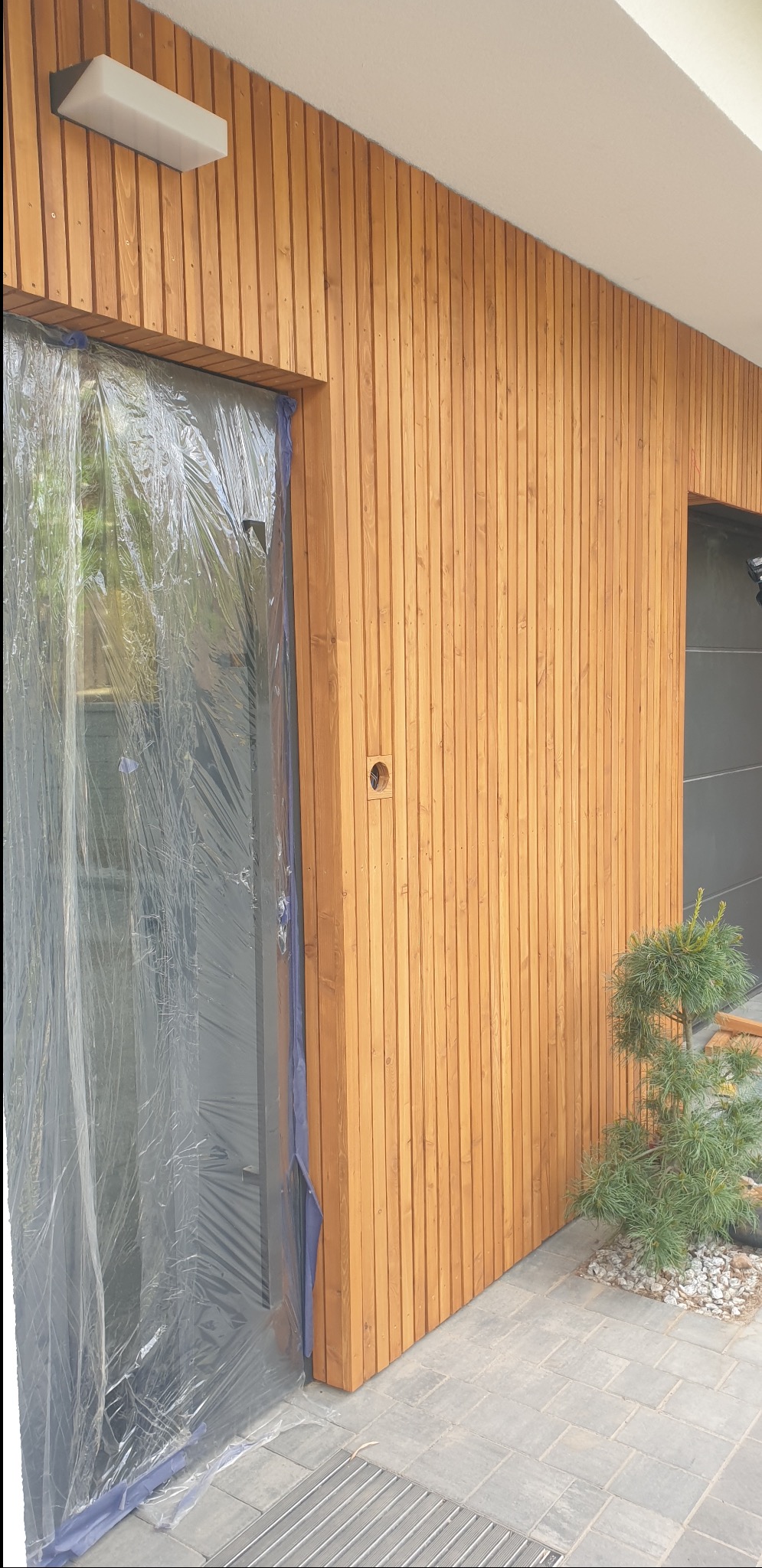 Nowoczesna elewacja budynku wykończona jasnym drewnem. Okno zabezpieczone folią malarską. Widoczne oświetlenie zewnętrzne i fragment chodnika.