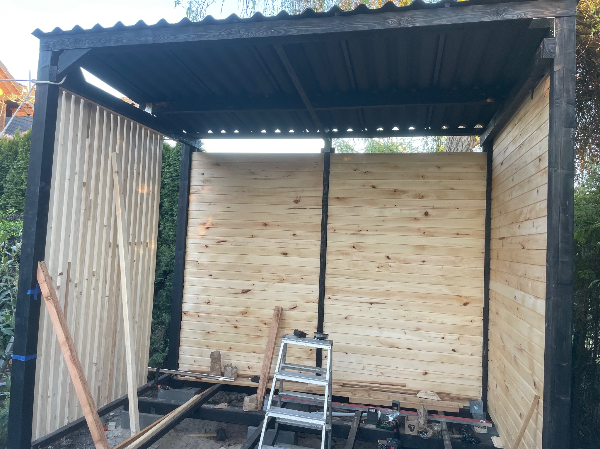 Budowa altany z jasnego drewna, widoczne ściany w trakcie montażu, czarne elementy konstrukcyjne. Na podłodze narzędzia i drabina.