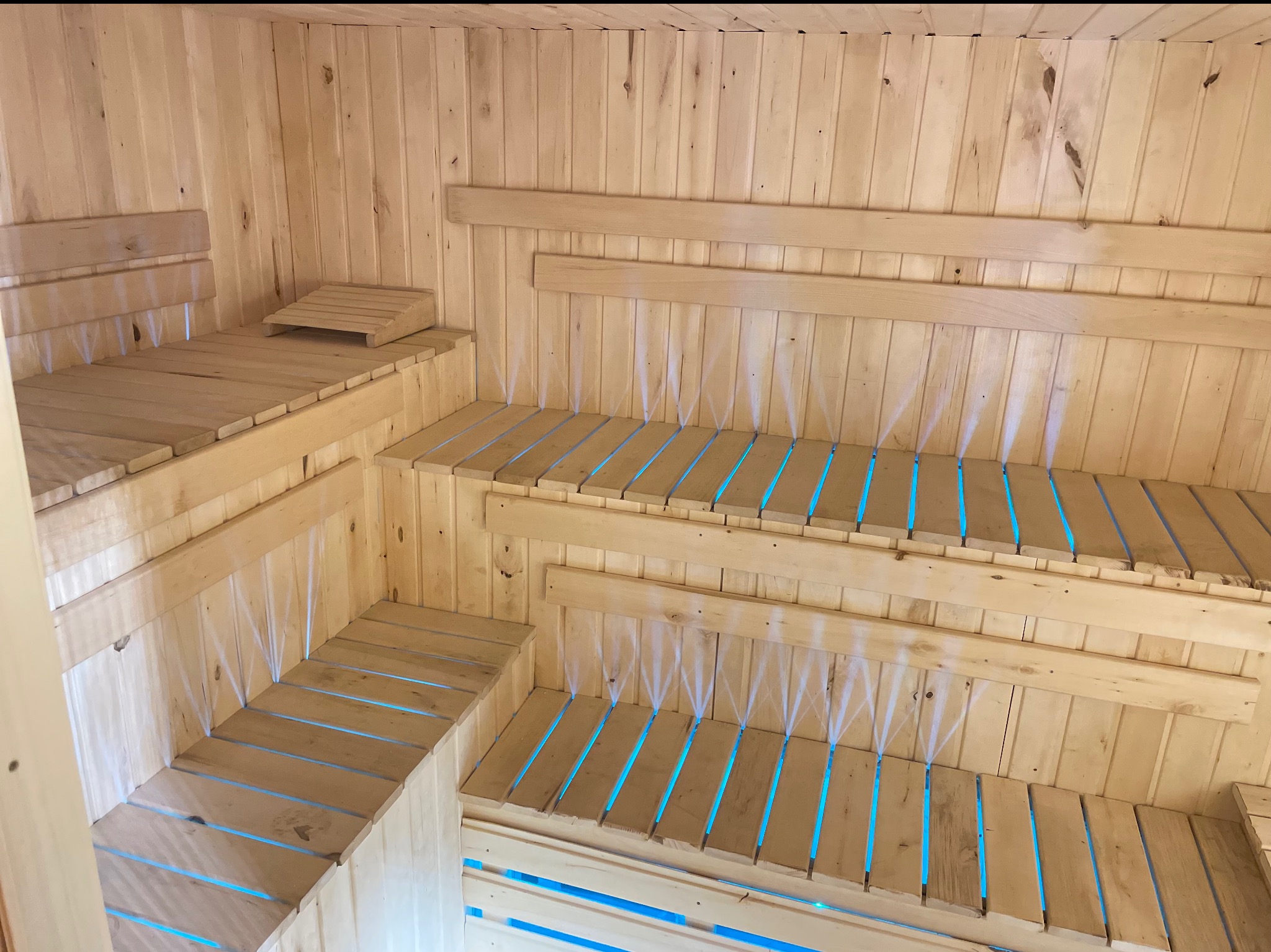 Jasne drewno i niebieskie podświetlenie wewnątrz sauny. Wielopoziomowe ławy z szczelinami, pionowe deski na ścianach i suficie, ciepłe światło.