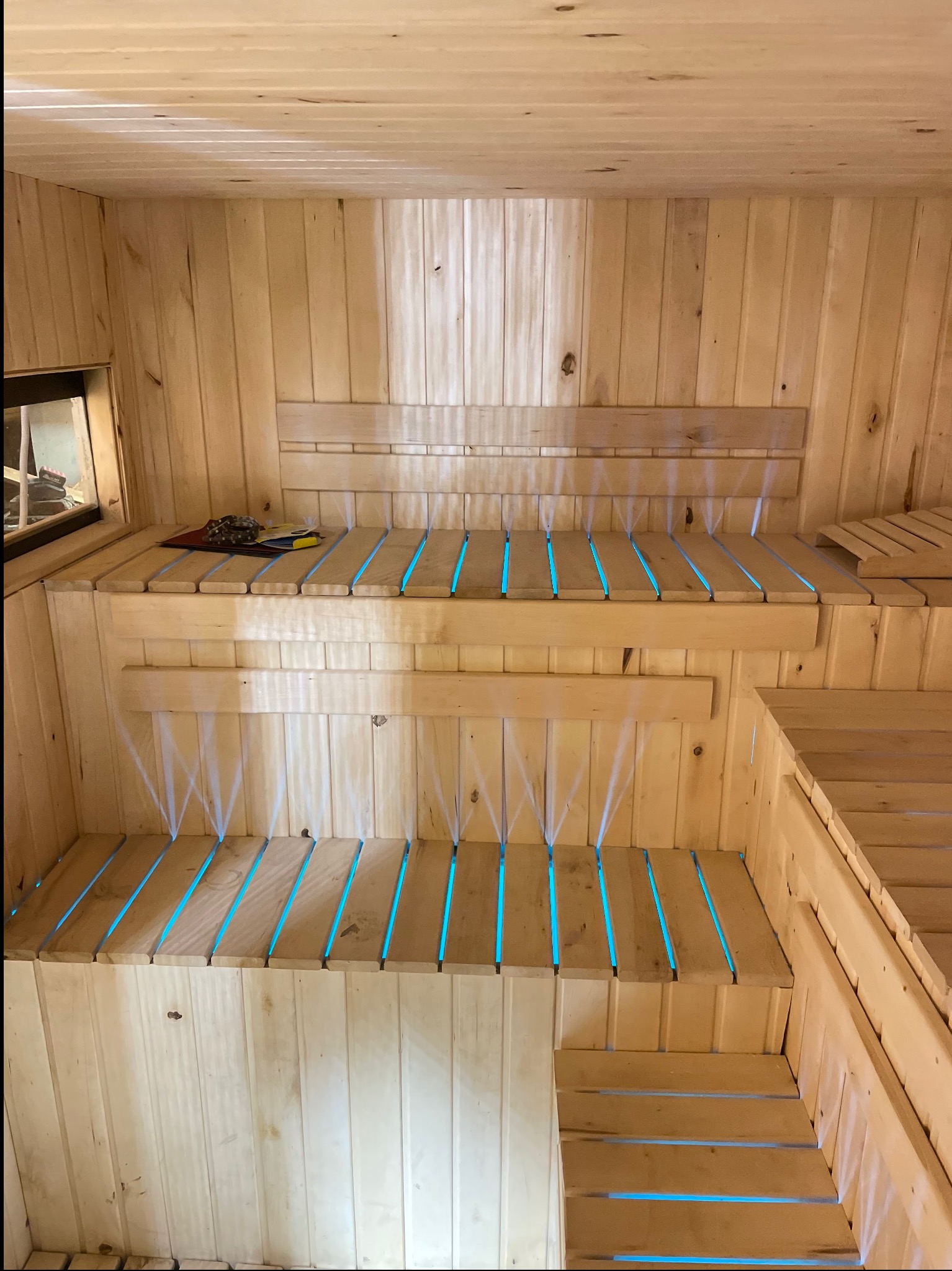 Wnętrze sauny z drewnianymi ławami, podświetlonymi na niebiesko, tworzącymi relaksującą atmosferę. Widoczne detale konstrukcyjne i narzędzia na półce.