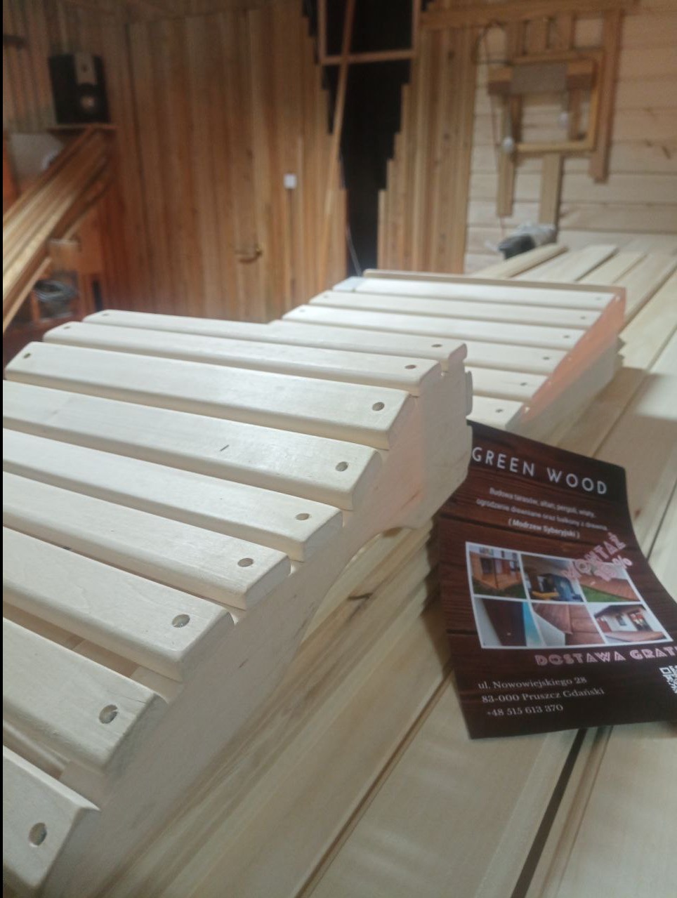 Fragment konstrukcji z jasnego drewna z nawierconymi otworami, na tle drewnianej ściany i ulotki firmy 'Green Wood' z adresem i numerem telefonu.