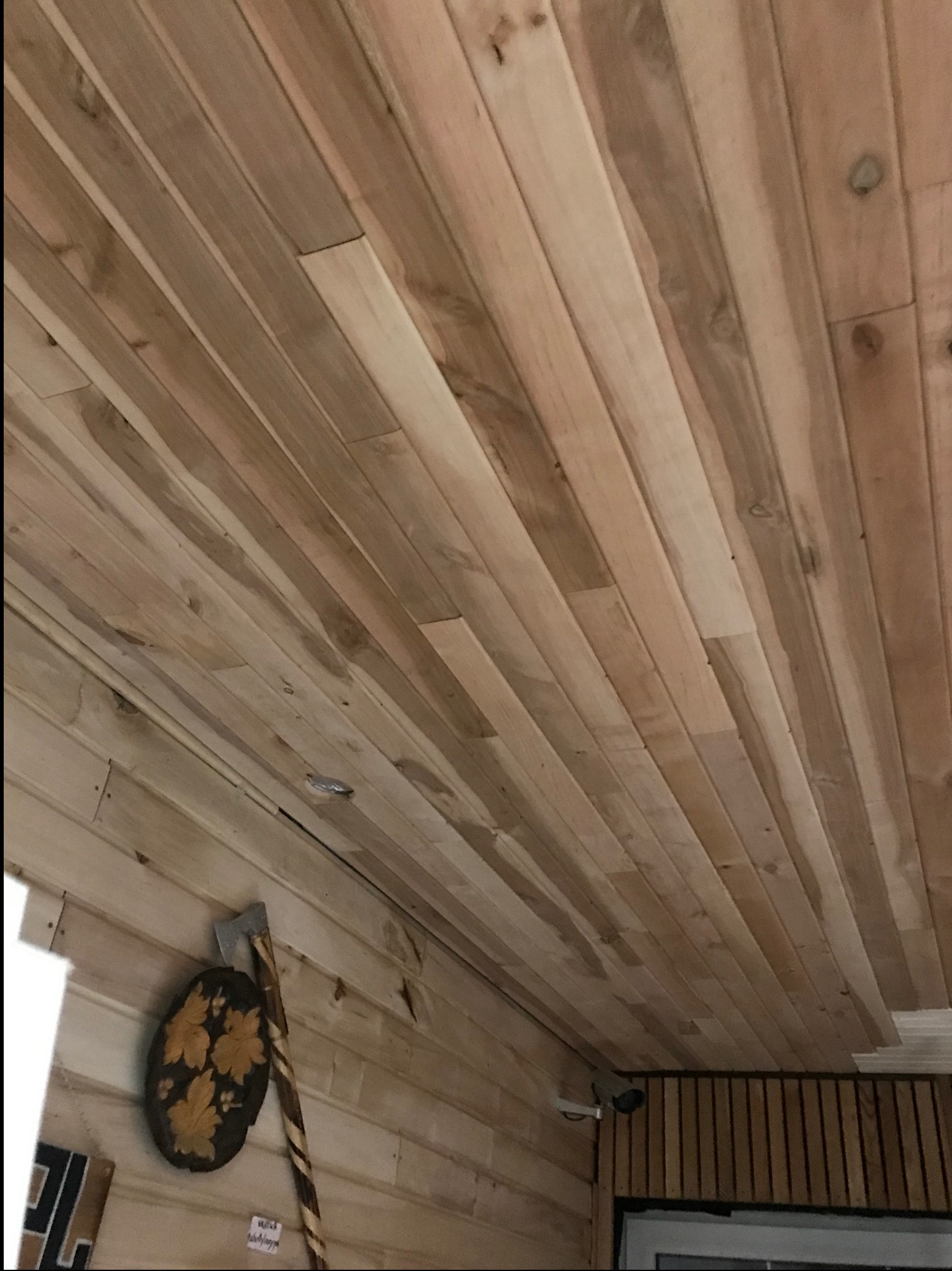 Wykończone drewnianą boazerią wnętrze sauny z widoczną kamerą monitoringu i dekoracyjnym toporem na ścianie. Jasne drewno, ukośne ułożenie desek.