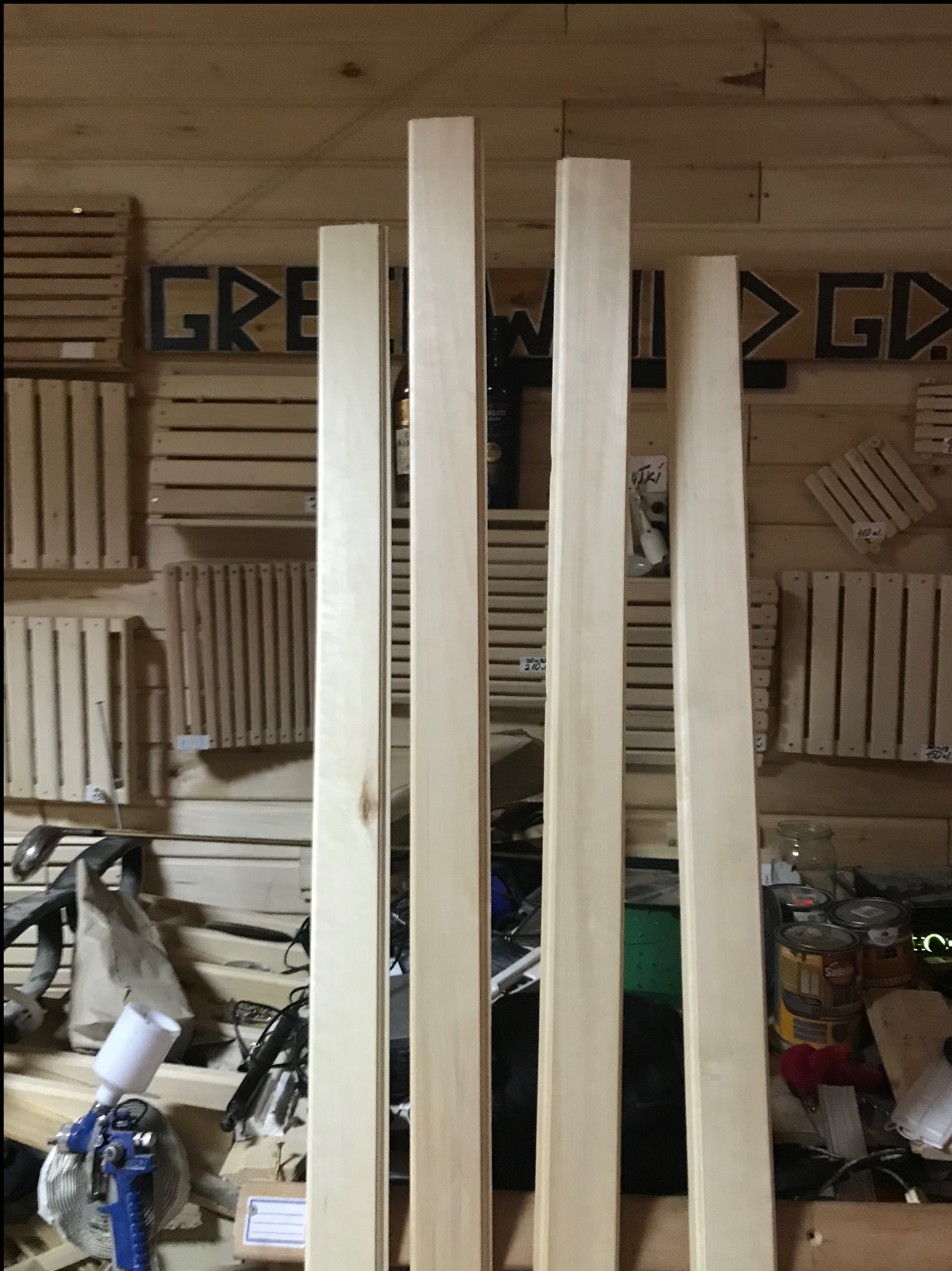 Cztery kantówki z jasnego drewna stoją pionowo na tle warsztatu z narzędziami i pojemnikami. Widoczny pistolet lakierniczy i puszki z farbą. Ściany warsztatu wyłożone drewnem.