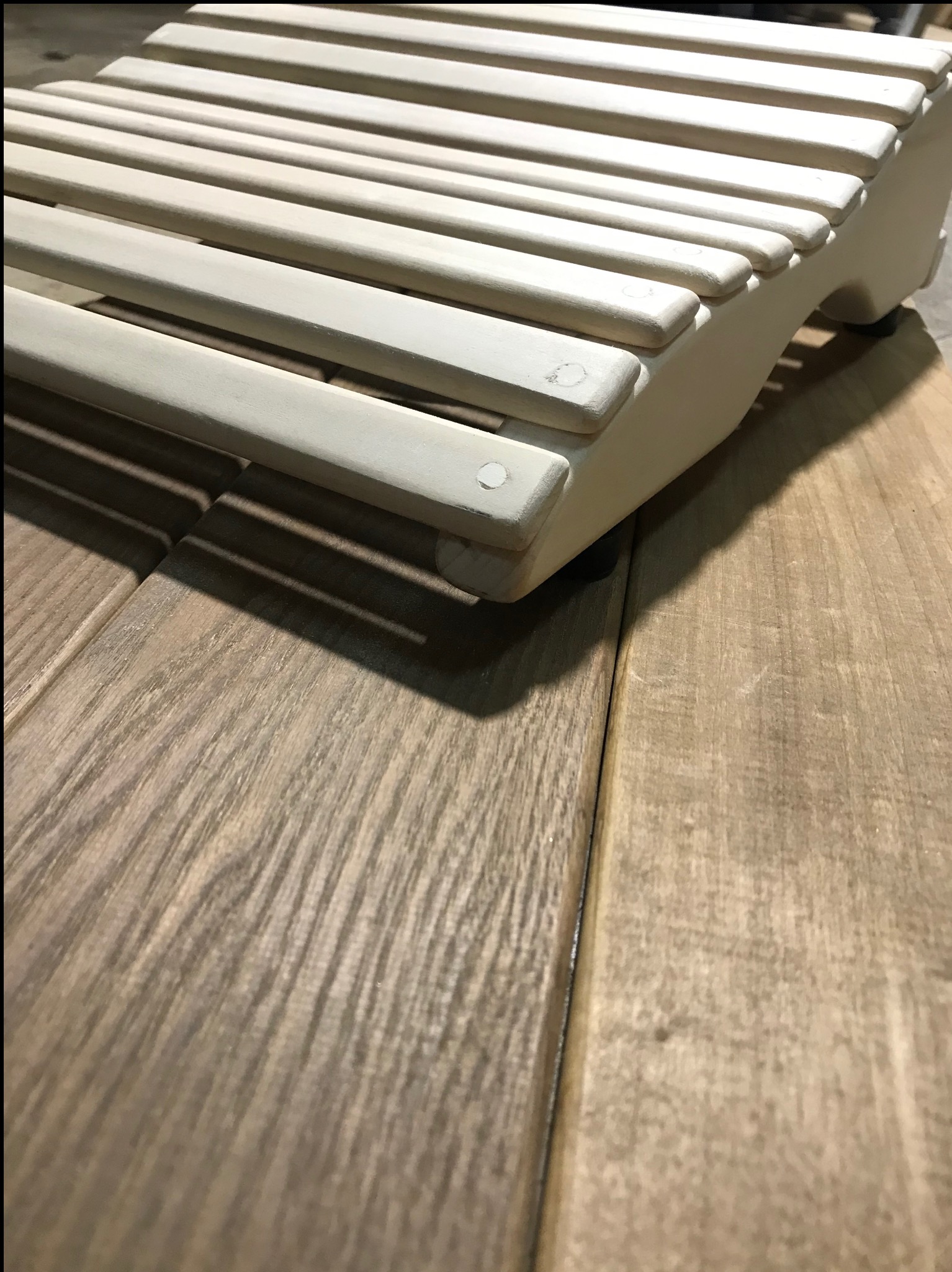 Biały, drewniany podnóżek z zaokrąglonymi szczebelkami, umieszczony na podłodze z paneli imitujących naturalne drewno. Widoczne łączenia i faktura drewna.