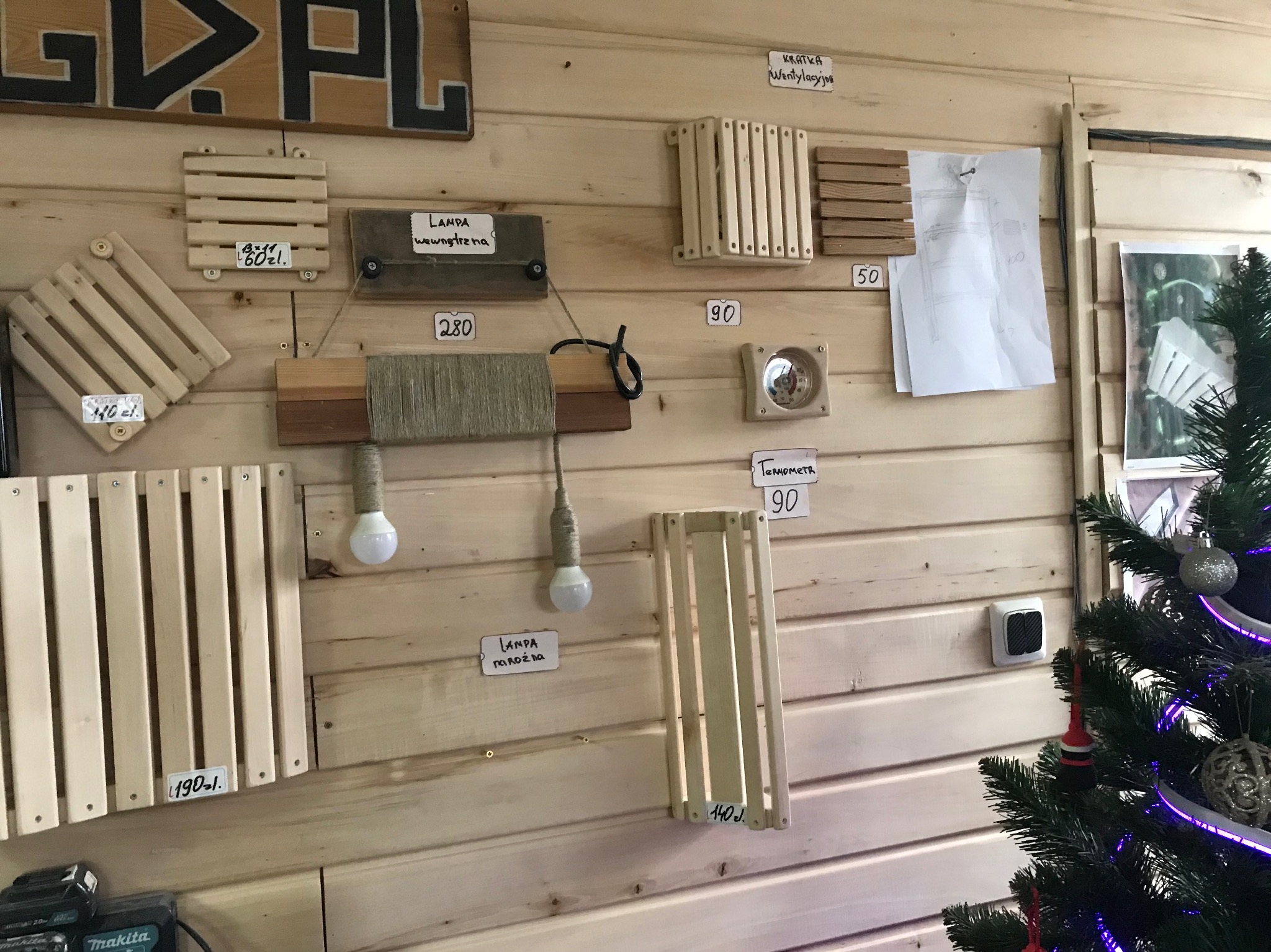 Ekspozycja drewnianych elementów: kratki wentylacyjne, lampy i termometr na ścianie z boazerii. Widoczne ceny produktów i częściowo choinka.