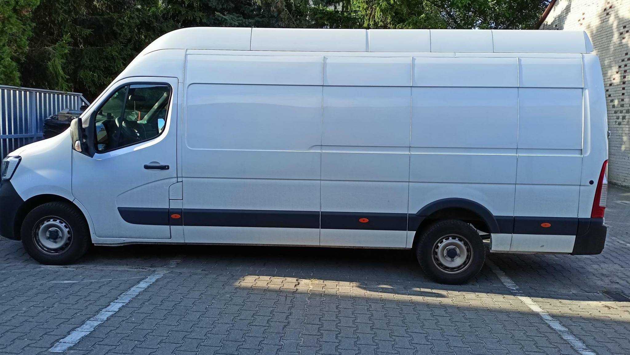 Biały, dostawczy bus Renault Master, zaparkowany na kostce brukowej, widok z boku, zarys drzew w tle, czyste, nieoklejone nadwozie.