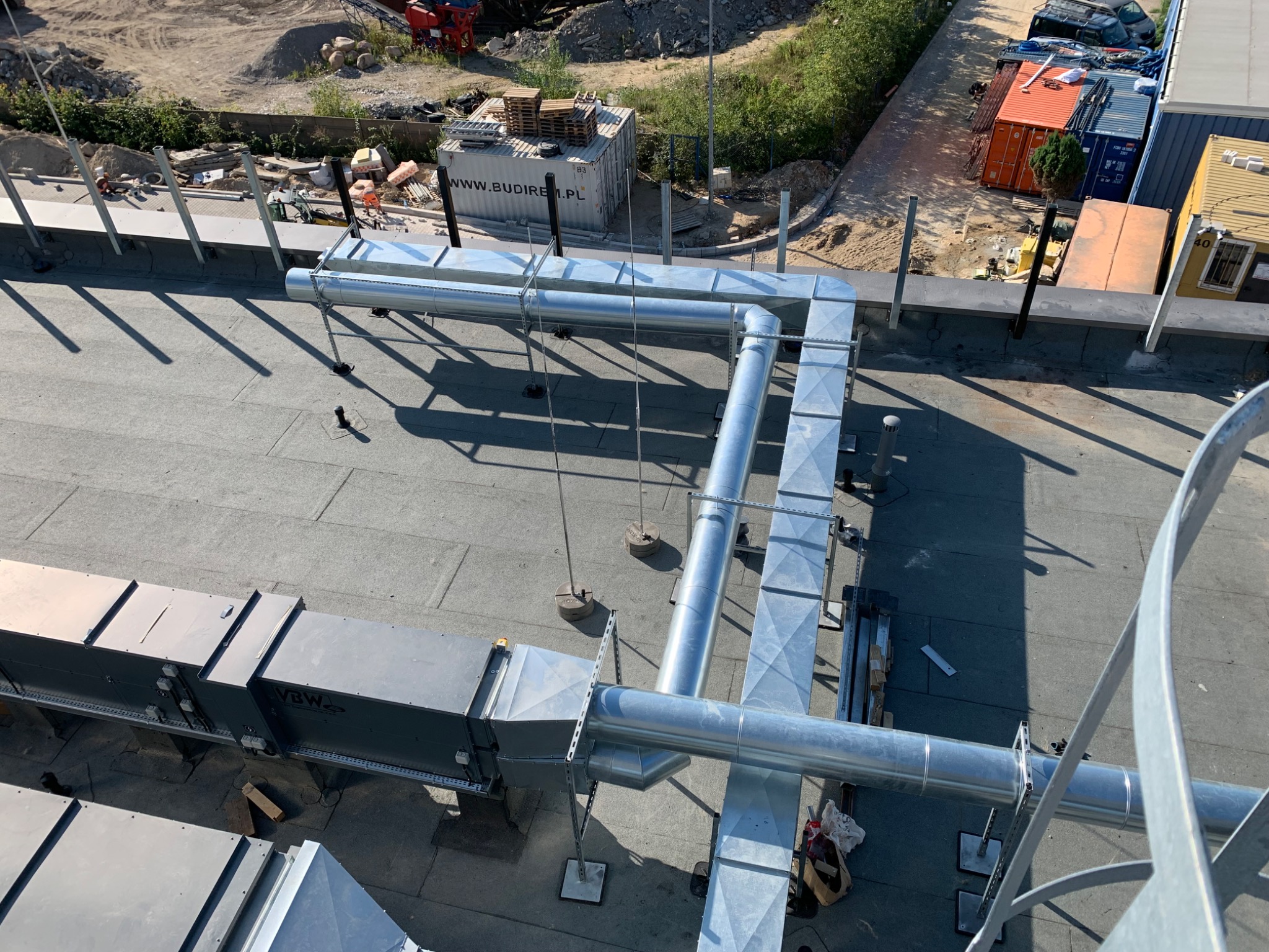 Widok z góry na zainstalowaną, srebrną instalację wentylacyjną na płaskim dachu budynku z widocznymi elementami konstrukcyjnymi i otoczeniem placu budowy.