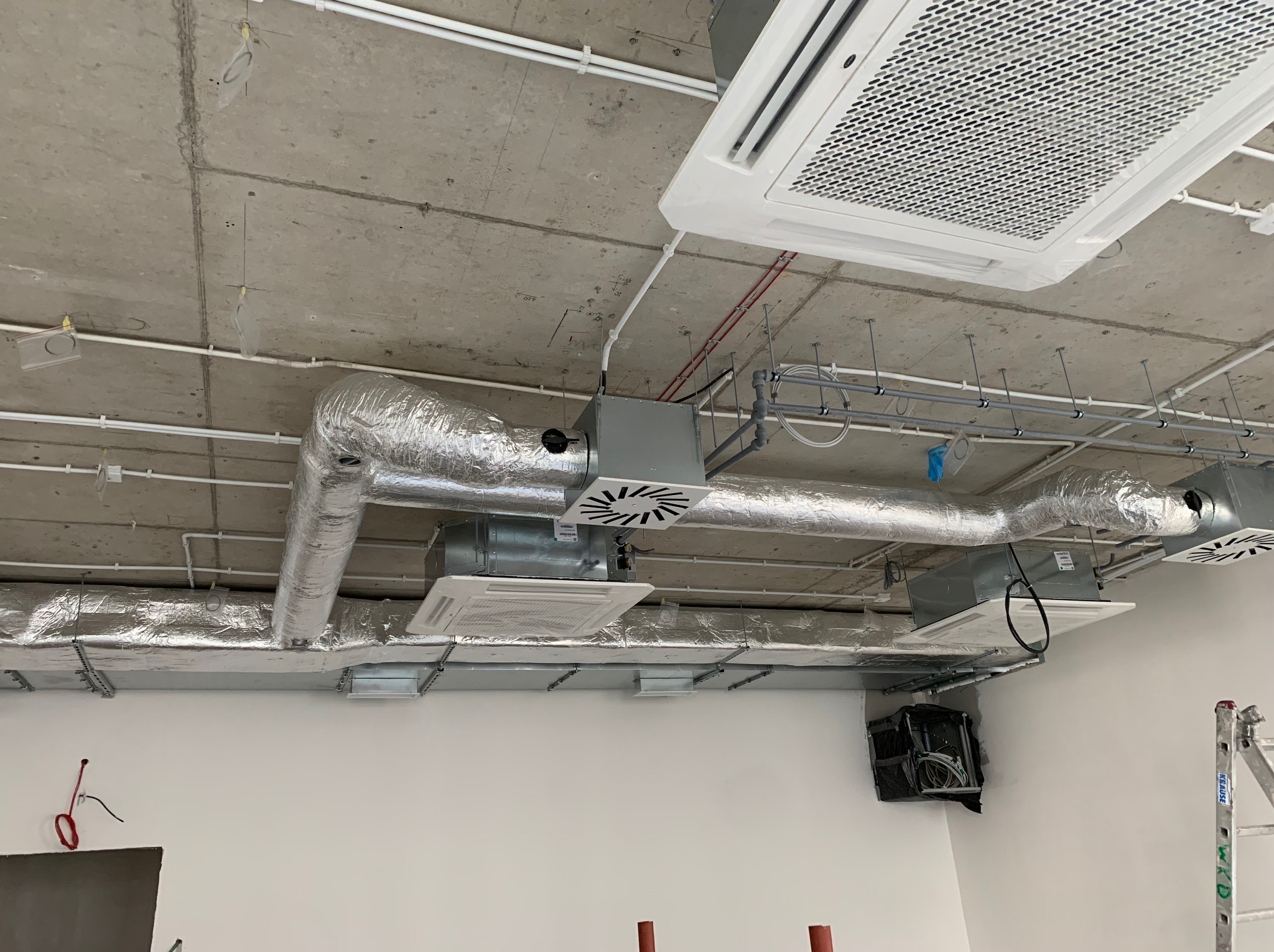 Sufit z widoczną instalacją wentylacyjną: kanały w srebrnej izolacji, białe rury i klimatyzatory kasetonowe. Surowy beton i fragment ściany w tle.