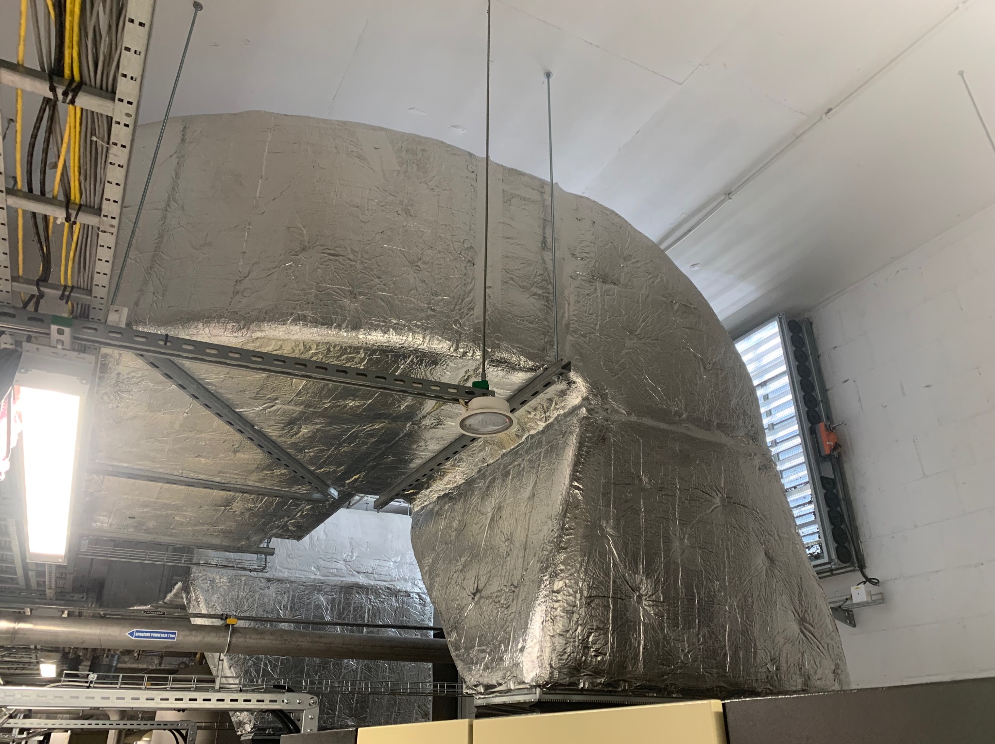 Izolowany kanał wentylacyjny z aluminium podwieszony pod sufitem, widoczne kable i elementy instalacji. Białe ściany i sufit. Oświetlenie LED.