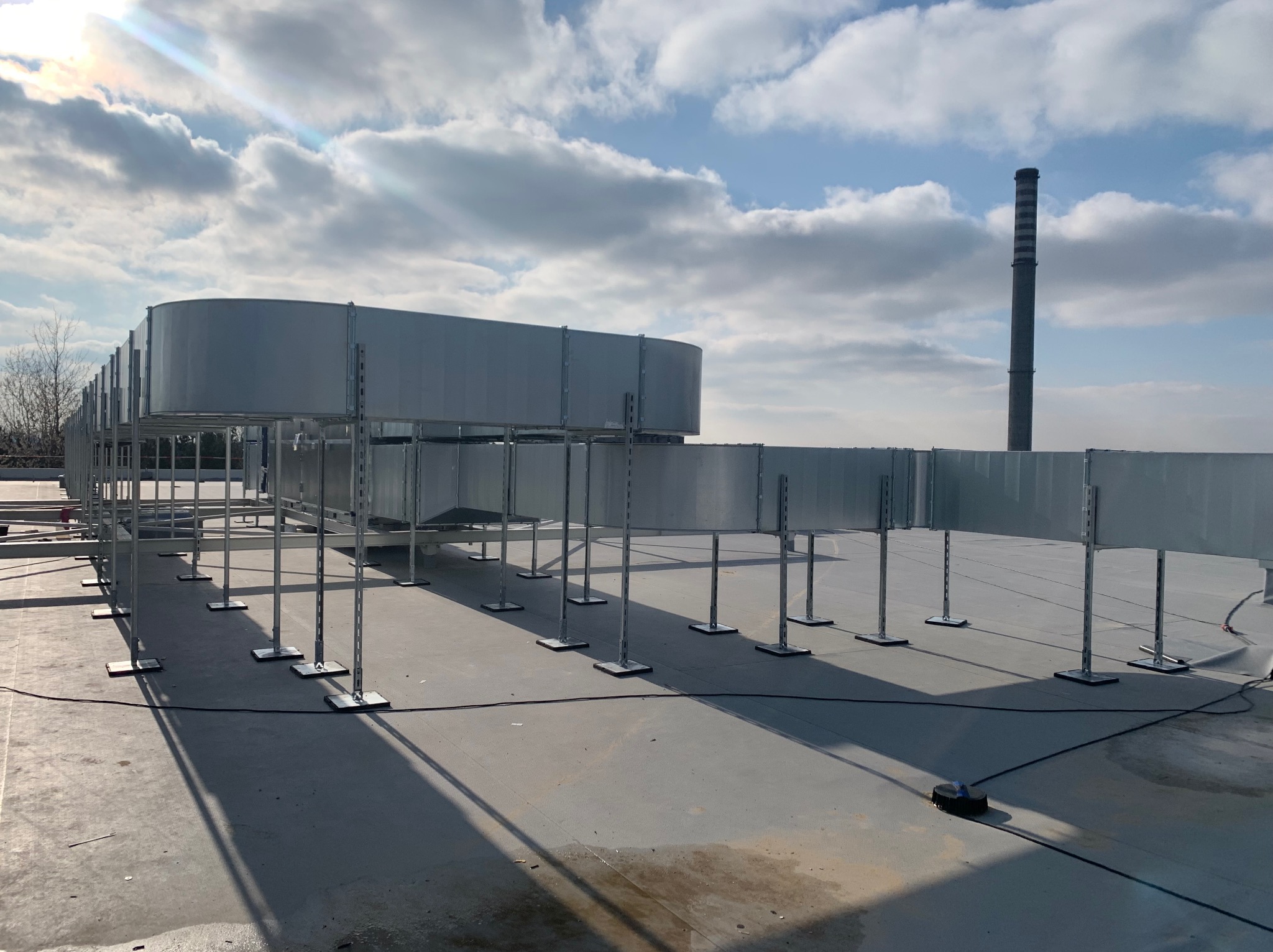 Montaż kanałów wentylacyjnych na dachu budynku komercyjnego, widoczny komin w tle, słoneczny dzień, niebieskie niebo z chmurami.