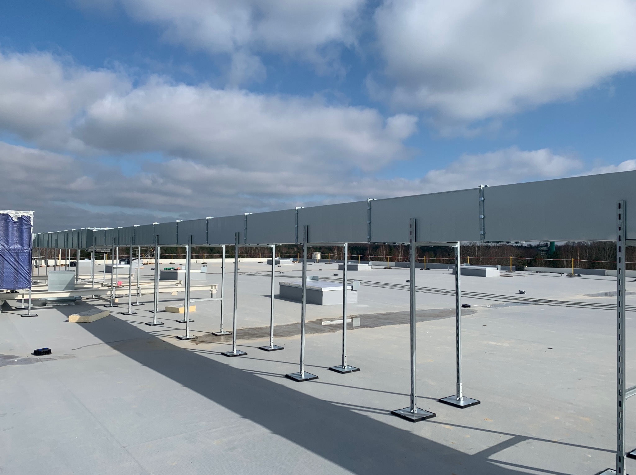 Montaż systemu wentylacji na dachu budynku komercyjnego. Widoczne wsporniki i elementy konstrukcyjne w trakcie instalacji, na tle błękitnego nieba z chmurami.