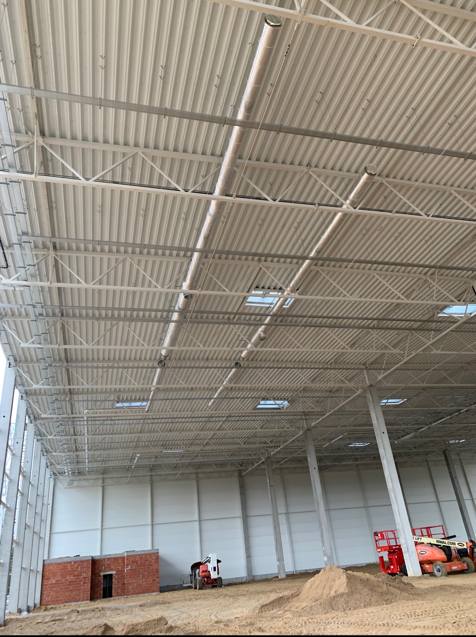 Wnętrze hali w budowie. Widoczne stalowe elementy konstrukcyjne dachu, rury wentylacyjne pod sufitem, ściany, podłoże oraz sprzęt budowlany.