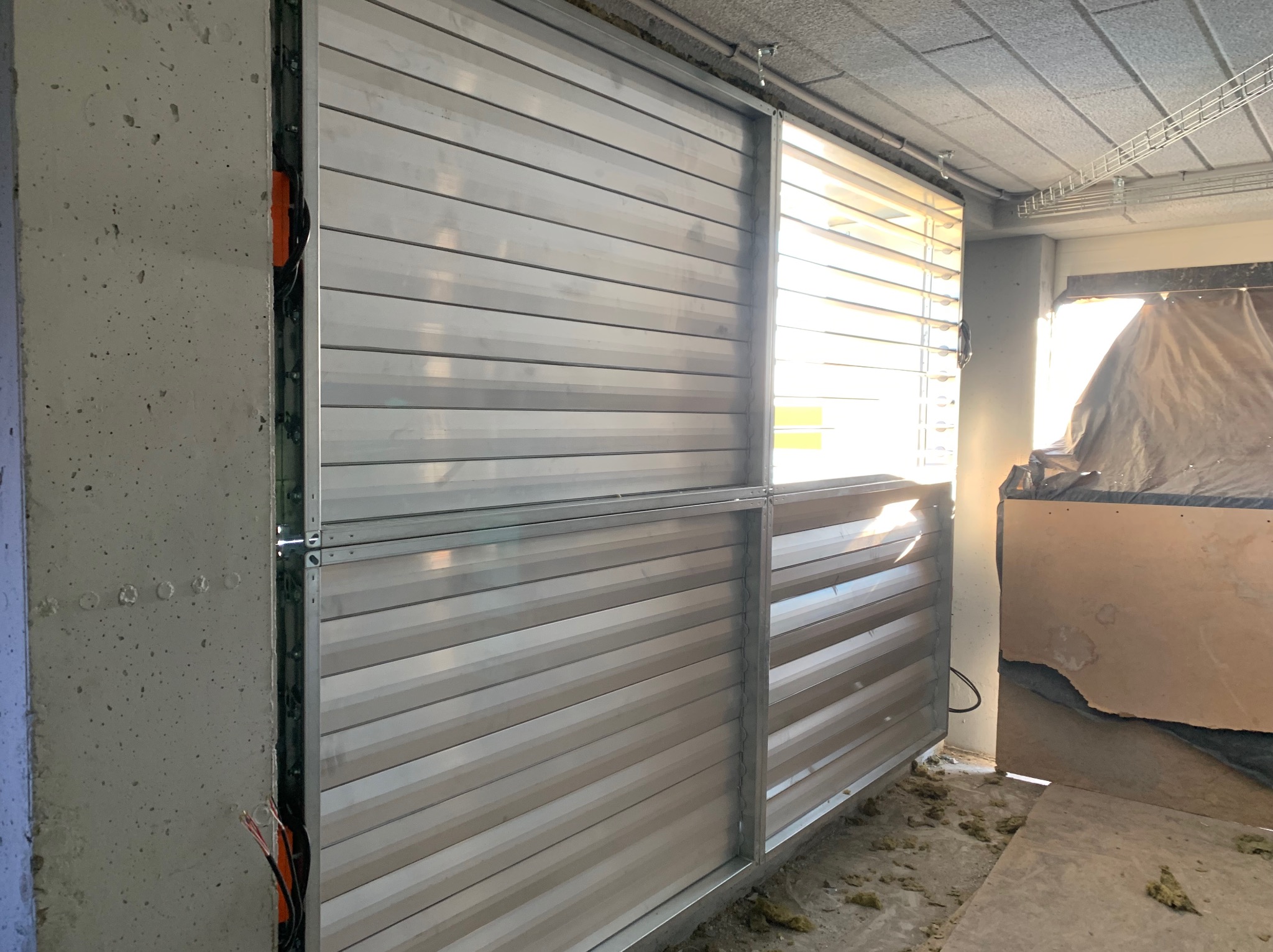 Szara, lamelowa obudowa wentylacyjna w surowym wnętrzu. Widoczne kable i fragmenty izolacji. Element systemu wentylacji w trakcie montażu.