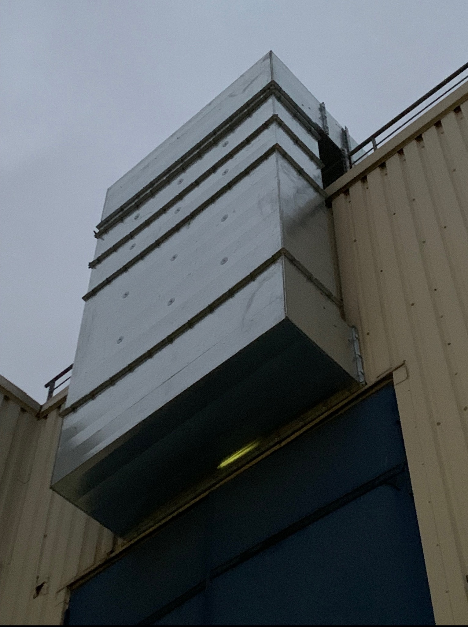 Zewnętrzny montaż systemu wentylacji, widoczny pionowy kanał wentylacyjny z blachy, przymocowany do ściany budynku. Ujęcie z dołu, pochmurne niebo w tle.