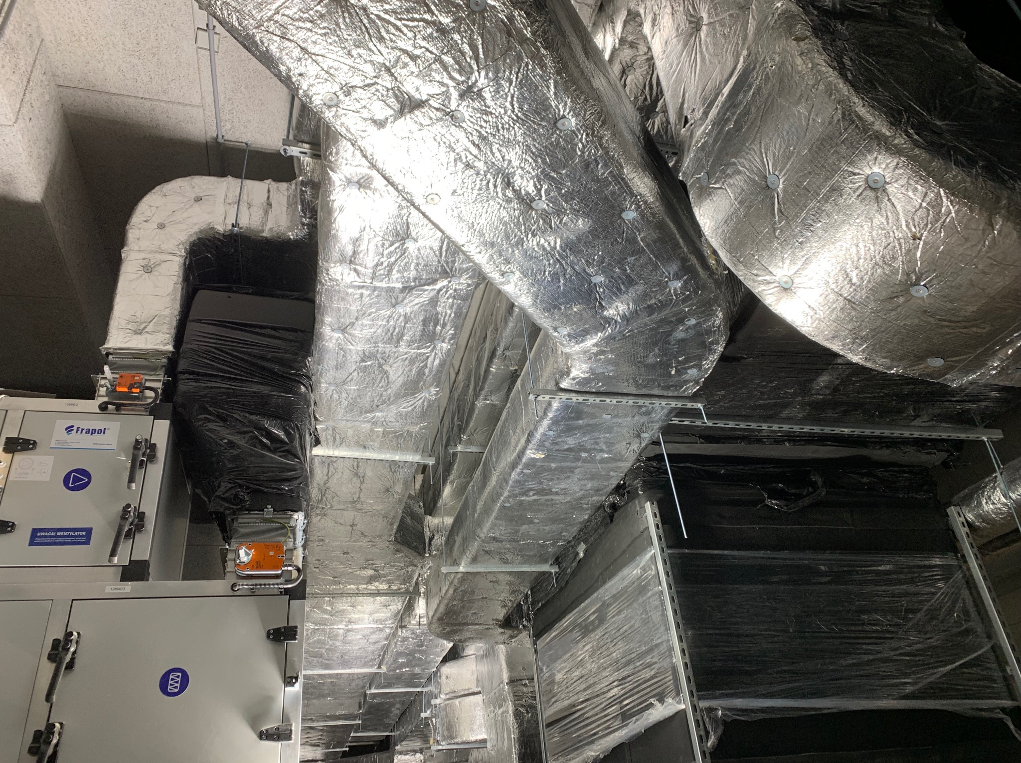 Kompleksowy system wentylacyjny z izolowanymi przewodami, widoczne elementy montażowe i jednostki centralne z etykietami informacyjnymi w pomieszczeniu technicznym.