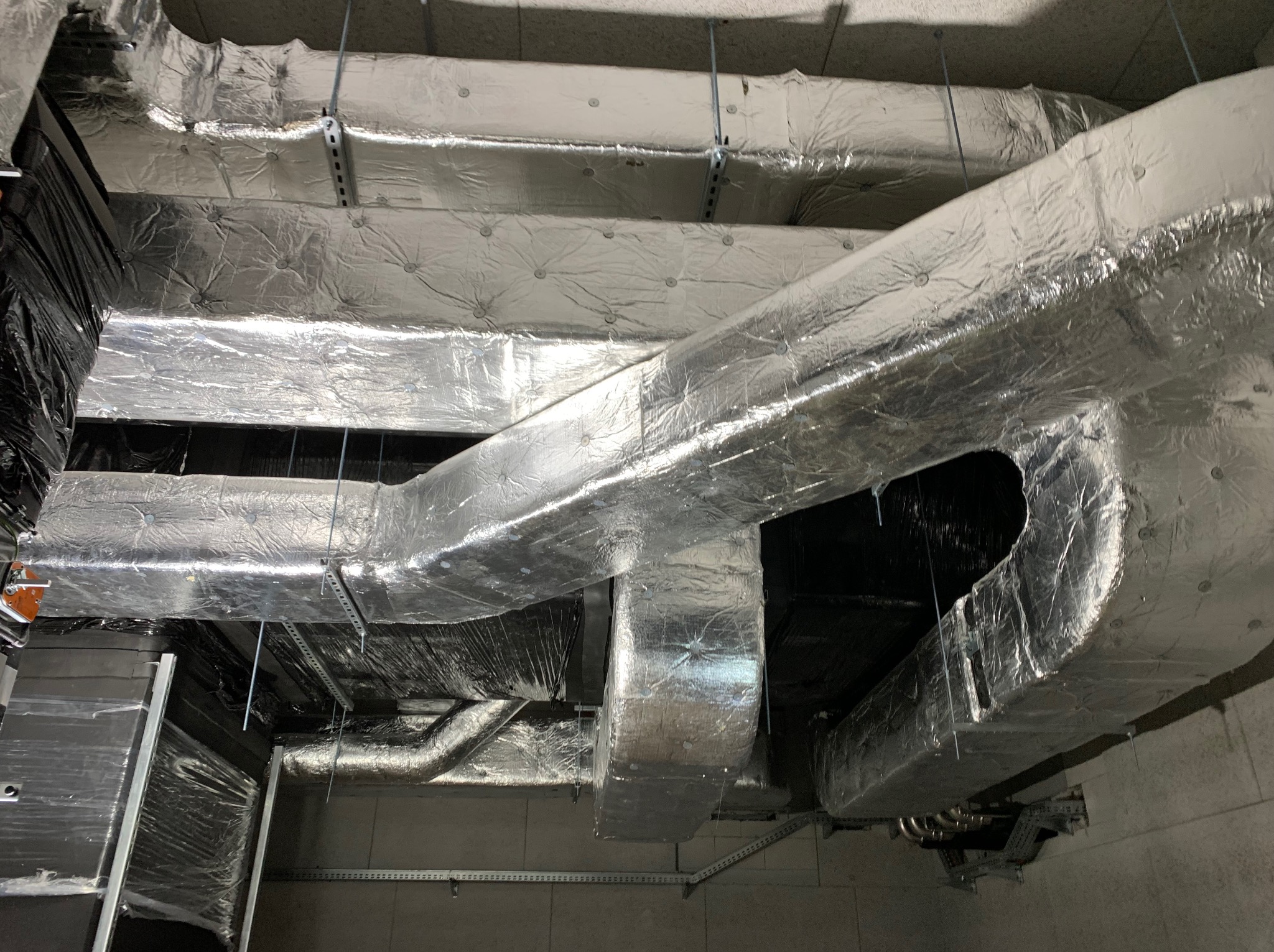 Izolowane kanały wentylacyjne zamontowane pod sufitem, widoczne elementy mocujące i fragment ściany. Instalacja w budynku komercyjnym lub przemysłowym.