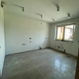 Jasne wnętrze po remoncie: szare płytki podłogowe, świeżo pomalowane białe ściany z gniazdkami i oknem. Widoczne lampy sufitowe.