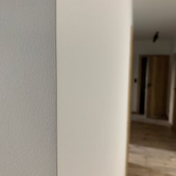 B&B Sylwia i Jacek Bednarek Sp. z o.o. - Fragment ściany po szpachlowaniu i malowaniu, widoczna faktura. W tle widok na wykończone wnętrze z drewnianą podłogą i drzwiami.