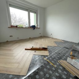 B&B Sylwia i Jacek Bednarek Sp. z o.o. - Układanie paneli podłogowych w jodełkę w jasnym pokoju. Widoczne narzędzia i podkład. Częściowo ułożona podłoga, okno z widokiem na drzewa.