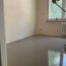 B&B Sylwia i Jacek Bednarek Sp. z o.o. - Wylewka samopoziomująca w pokoju, świeżo rozprowadzona. W tle okno z widokiem na zieleń i fragment grzejnika. Widoczne narzędzie do rozprowadzania wylewki.