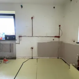 Przygotowanie do ogrzewania podłogowego: ułożona izolacja termiczna i instalacja elektryczna w ścianie, widoczne puszki i przewody. Częściowo zagruntowana ściana.