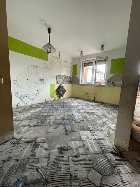 Demontaż starej podłogi z płytek w trakcie remontu mieszkania. Widoczne fragmenty płytek i kleju, ściany z resztkami farby i tapet. Przygotowanie do dalszych prac wykończeniowych.