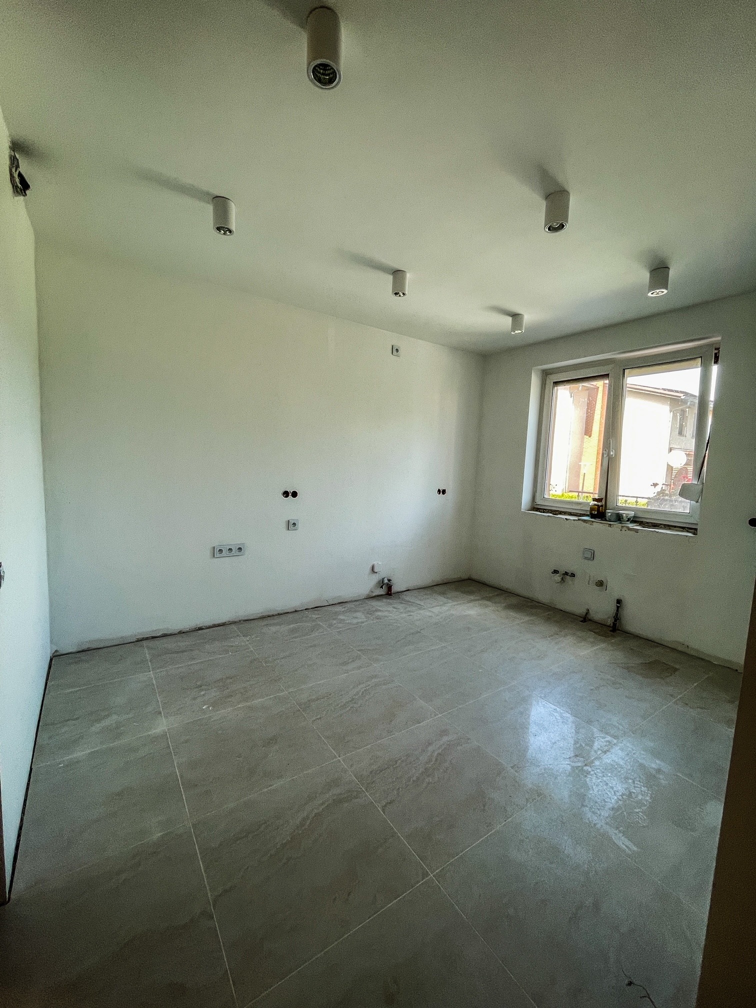 Jasne wnętrze po remoncie: szare płytki podłogowe, świeżo pomalowane białe ściany z gniazdkami i oknem. Widoczne lampy sufitowe.