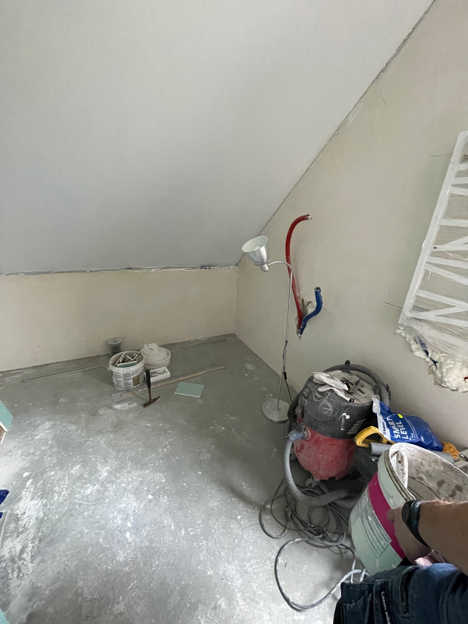 Pokój ze skośnym sufitem w trakcie prac przygotowawczych: szpachlowanie ścian, narzędzia, lampa i odkurzacz budowlany. Widok na wnętrze w trakcie remontu.