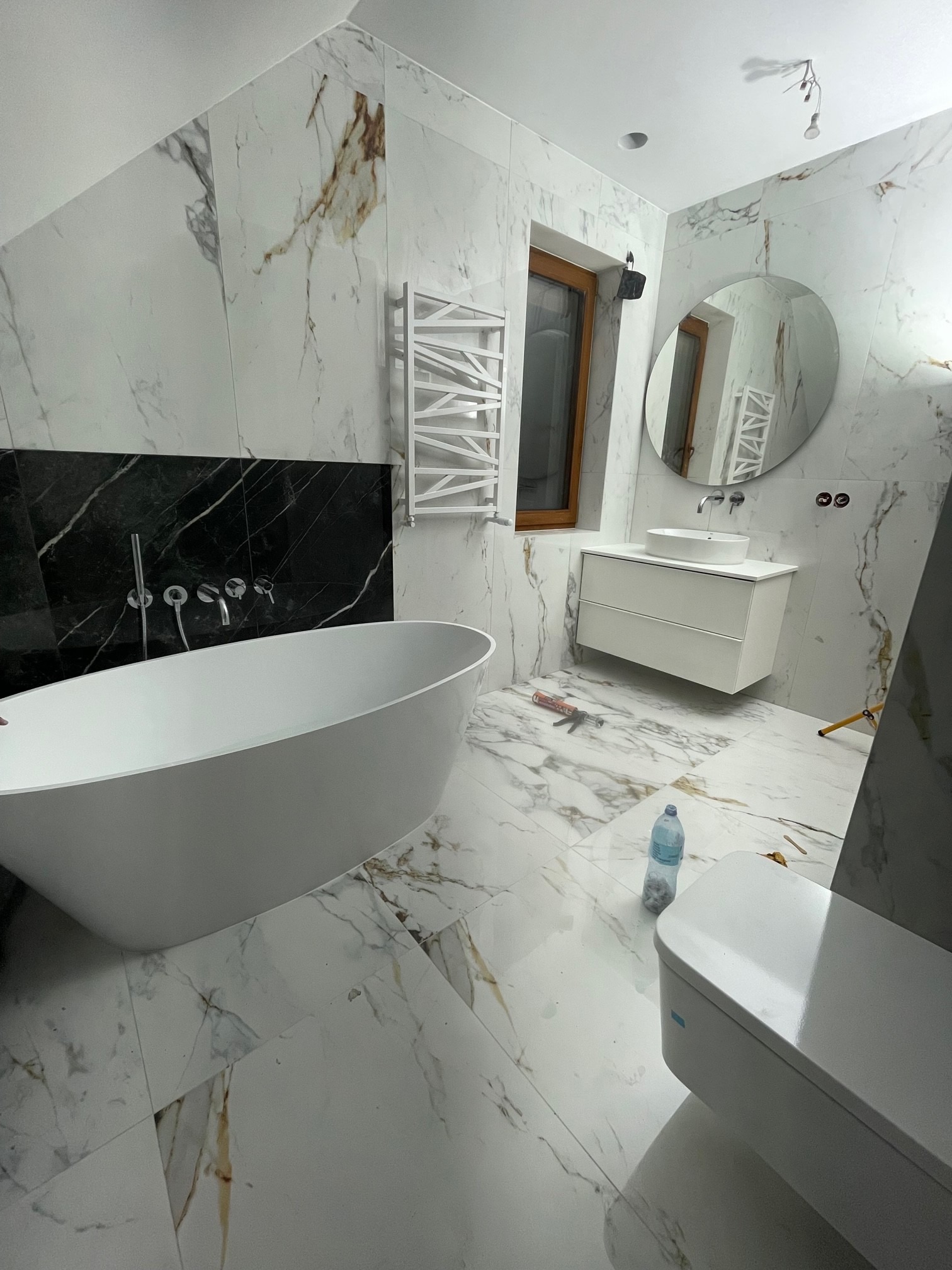 Elegancka łazienka z wanną wolnostojącą, marmurowymi płytkami i okrągłym lustrem. Widoczne narzędzia sugerują prace wykończeniowe.