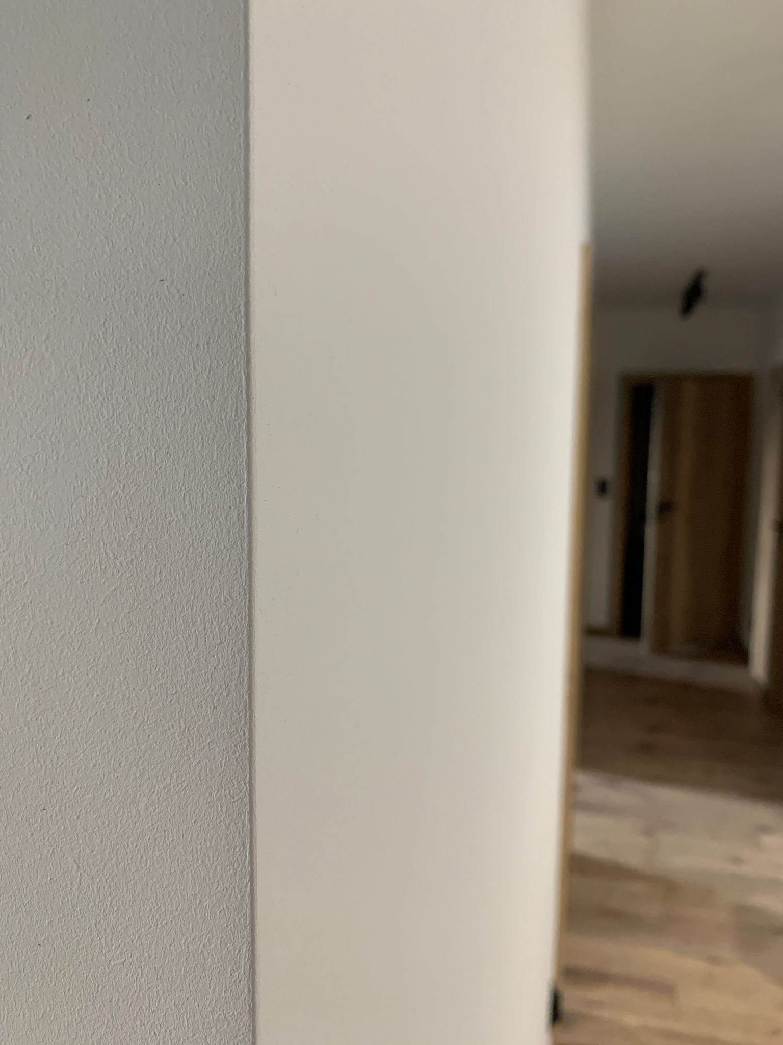 Fragment ściany po szpachlowaniu i malowaniu, widoczna faktura. W tle widok na wykończone wnętrze z drewnianą podłogą i drzwiami.