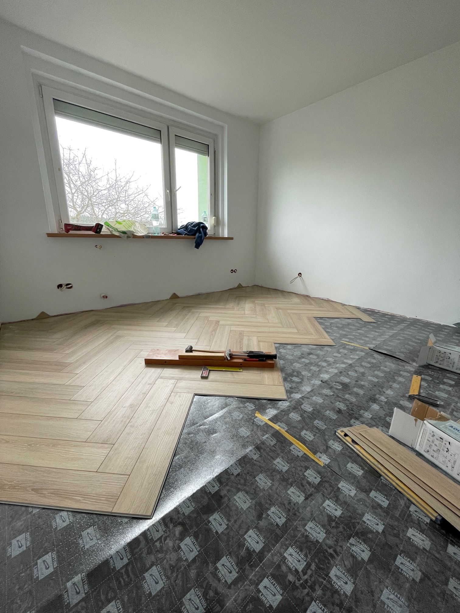 Układanie paneli podłogowych w jodełkę w jasnym pokoju. Widoczne narzędzia i podkład. Częściowo ułożona podłoga, okno z widokiem na drzewa.