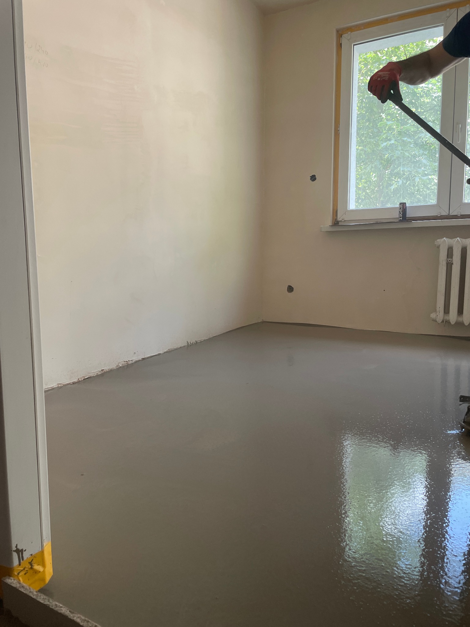 Wylewka samopoziomująca w pokoju, świeżo rozprowadzona. W tle okno z widokiem na zieleń i fragment grzejnika. Widoczne narzędzie do rozprowadzania wylewki.