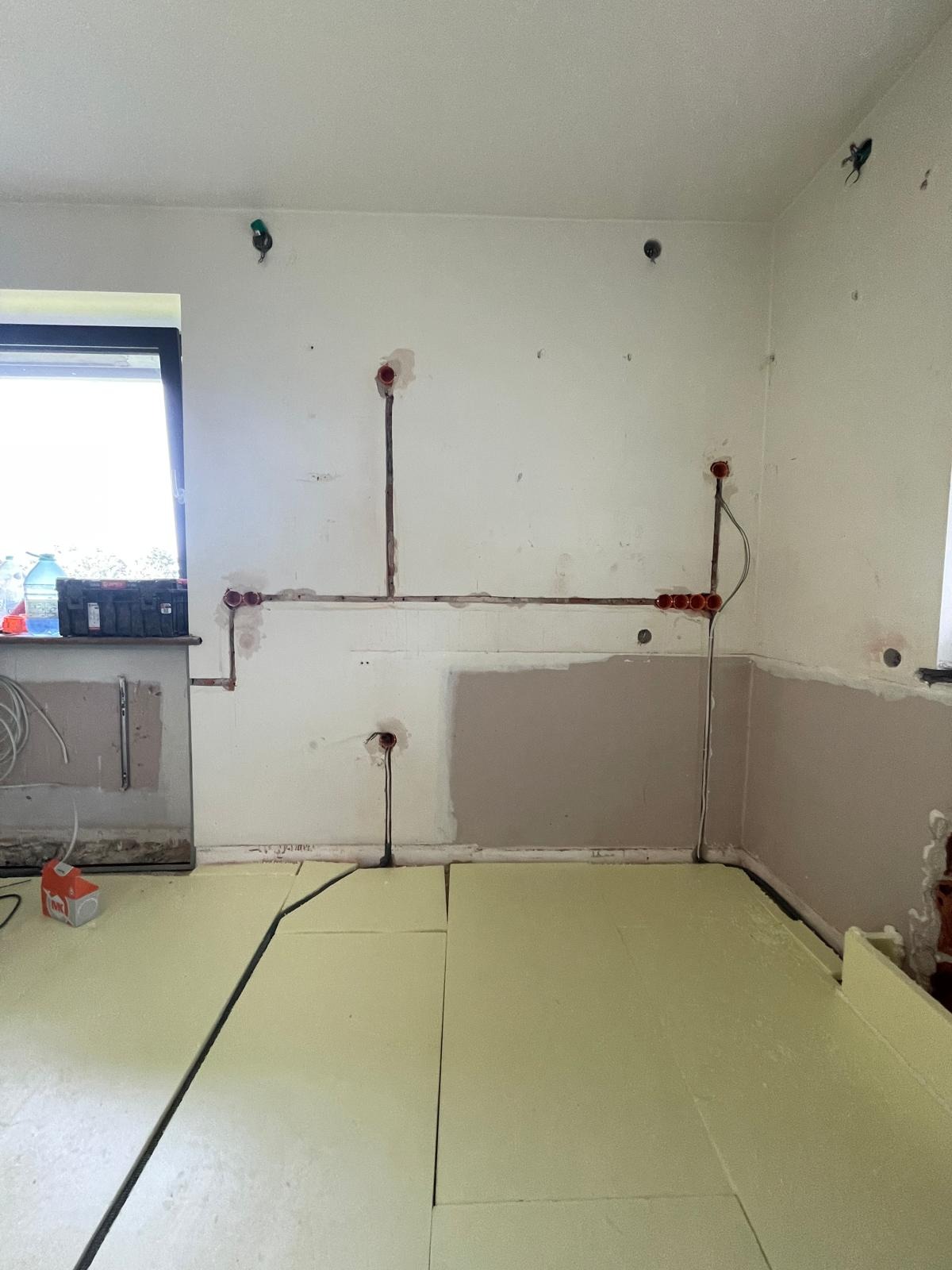 Przygotowanie do ogrzewania podłogowego: ułożona izolacja termiczna i instalacja elektryczna w ścianie, widoczne puszki i przewody. Częściowo zagruntowana ściana.