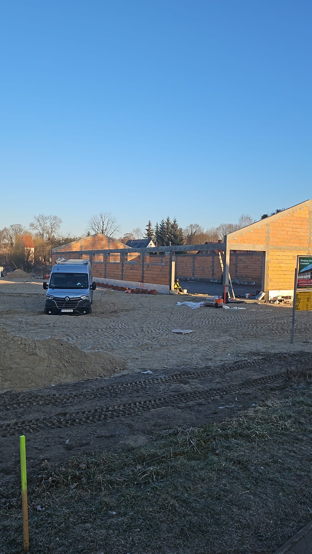 Budowa domu w trakcie wznoszenia ścian z czerwonej cegły, na placu budowy stoi srebrny bus. Widoczne narzędzia i pracownik w odblaskowej kamizelce.
