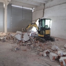 DK TRANS-KOP SP. Z O.O. - Żółta minikoparka Yanmar w trakcie rozbiórki muru wewnątrz hali. Gruz ceglany i betonowy na podłodze. Widoczne ślady pracy maszyny.