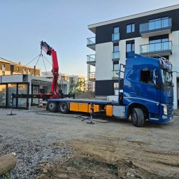 DK TRANS-KOP SP. Z O.O. - Niebieska ciężarówka z dźwigiem transportuje modułowy pawilon na placu budowy, w tle nowoczesne budynki mieszkalne i przenośna toaleta.