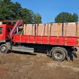 DK TRANS-KOP SP. Z O.O. - Czerwona ciężarówka z dźwigiem AMCO przewozi palety cegieł, zabezpieczone żółtymi pasami. W tle widoczny zielony kontener i mała koparka.