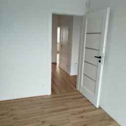 Jasne panele podłogowe w pokoju z białymi ścianami i otwartymi drzwiami, widok na sąsiednie pomieszczenie. Nowoczesne wykończenie wnętrza.