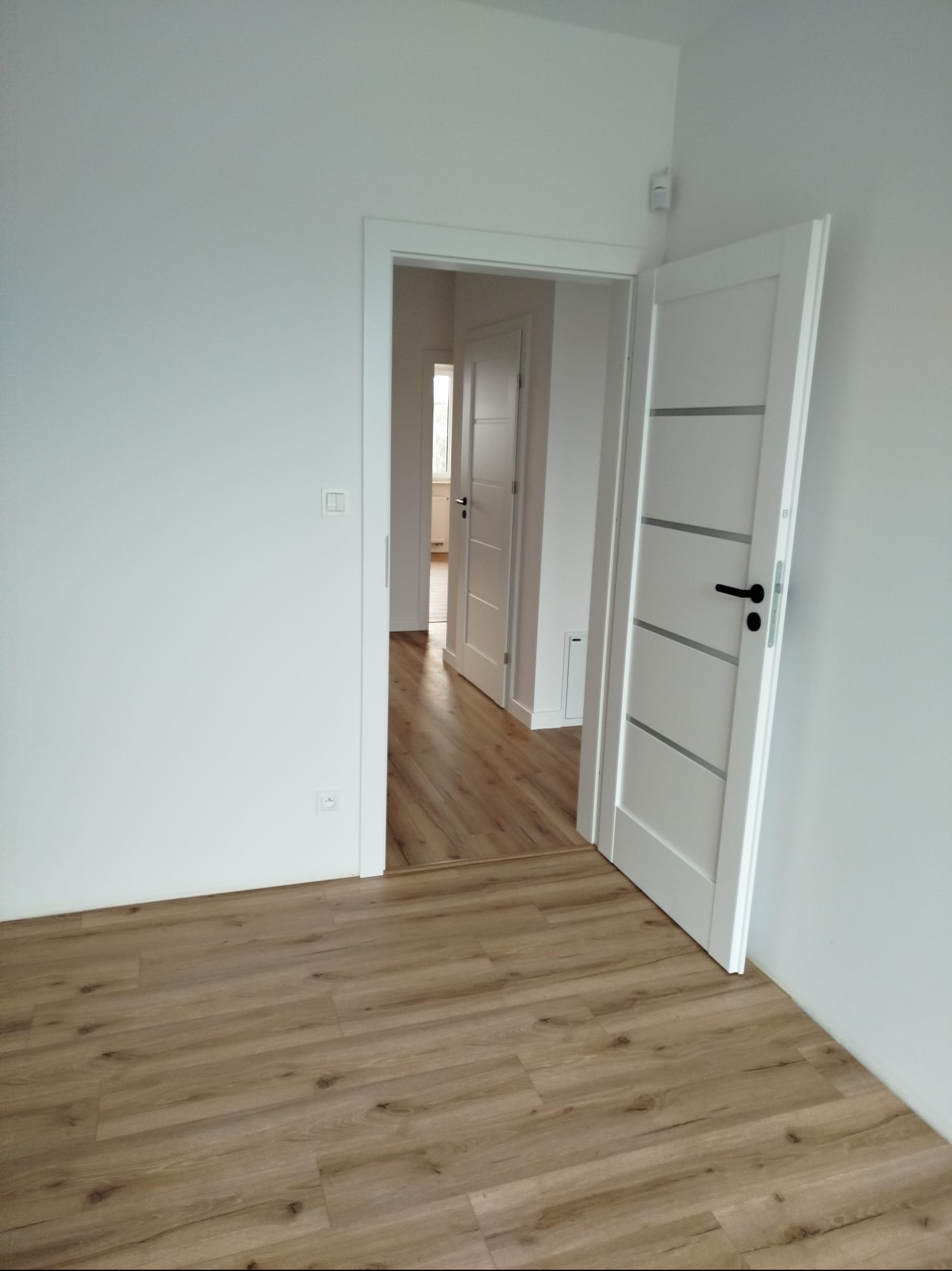 Jasne panele podłogowe w pokoju z białymi ścianami i otwartymi drzwiami. Widok na sąsiednie pomieszczenie z identycznym wykończeniem. Nowoczesny design.