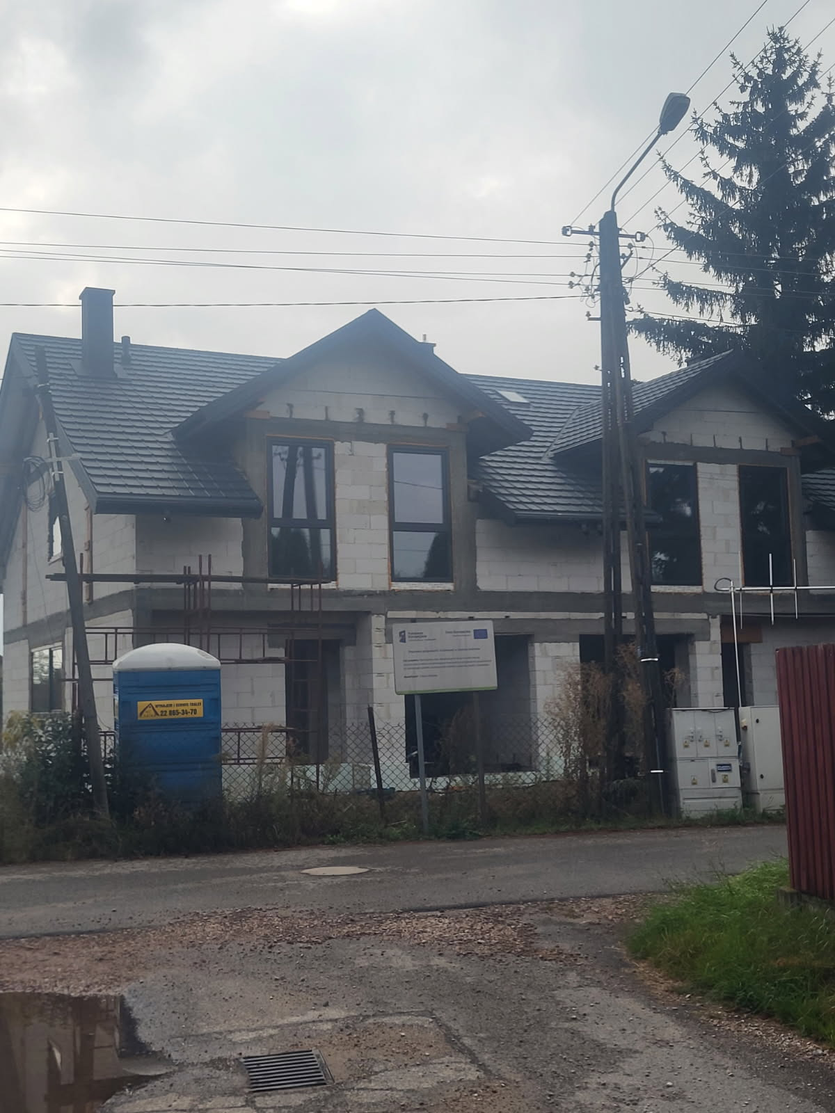 Surowy stan domu murowanego z ciemną dachówką, widoczne okna i rusztowania. Na pierwszym planie toaleta przenośna i tablica informacyjna. Pochmurne niebo w tle.