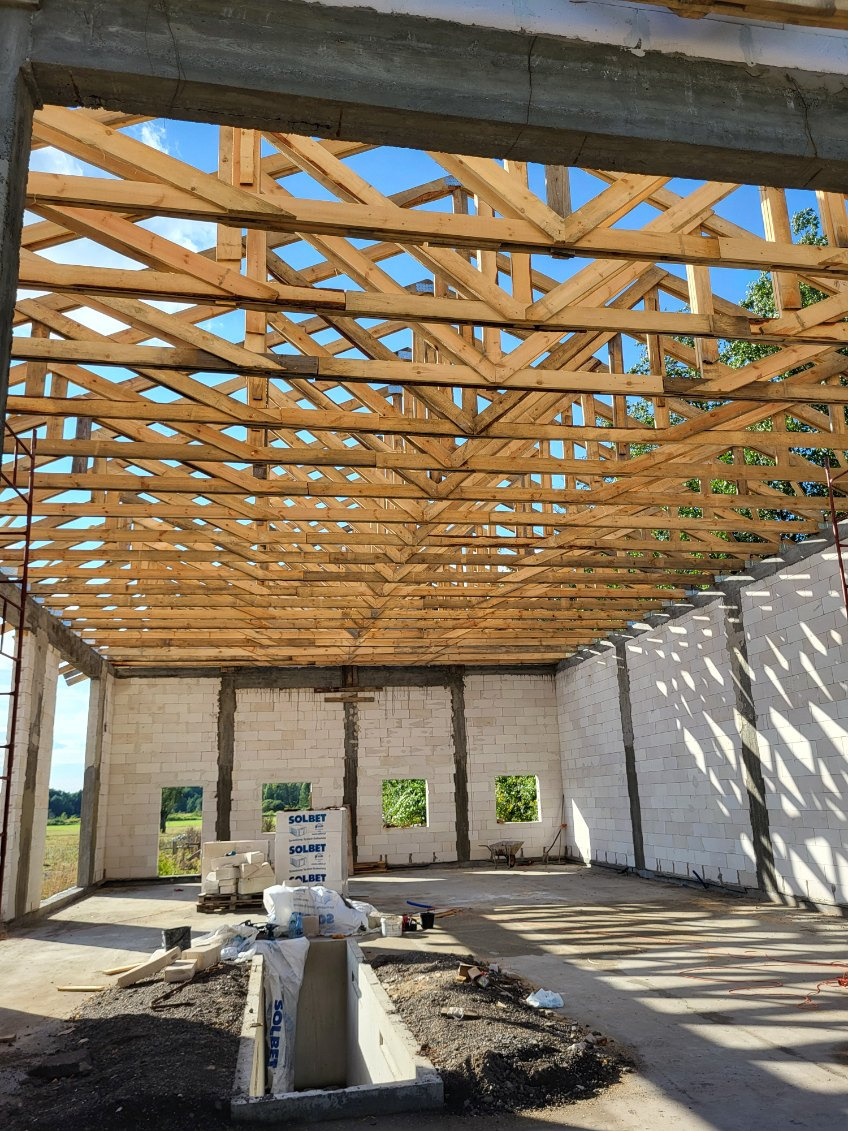 Wnętrze surowego domu z białego betonu komórkowego, widoczna drewniana konstrukcja dachu i palety z bloczkami Solbet. Budowa w toku, widok na zewnątrz przez okna.