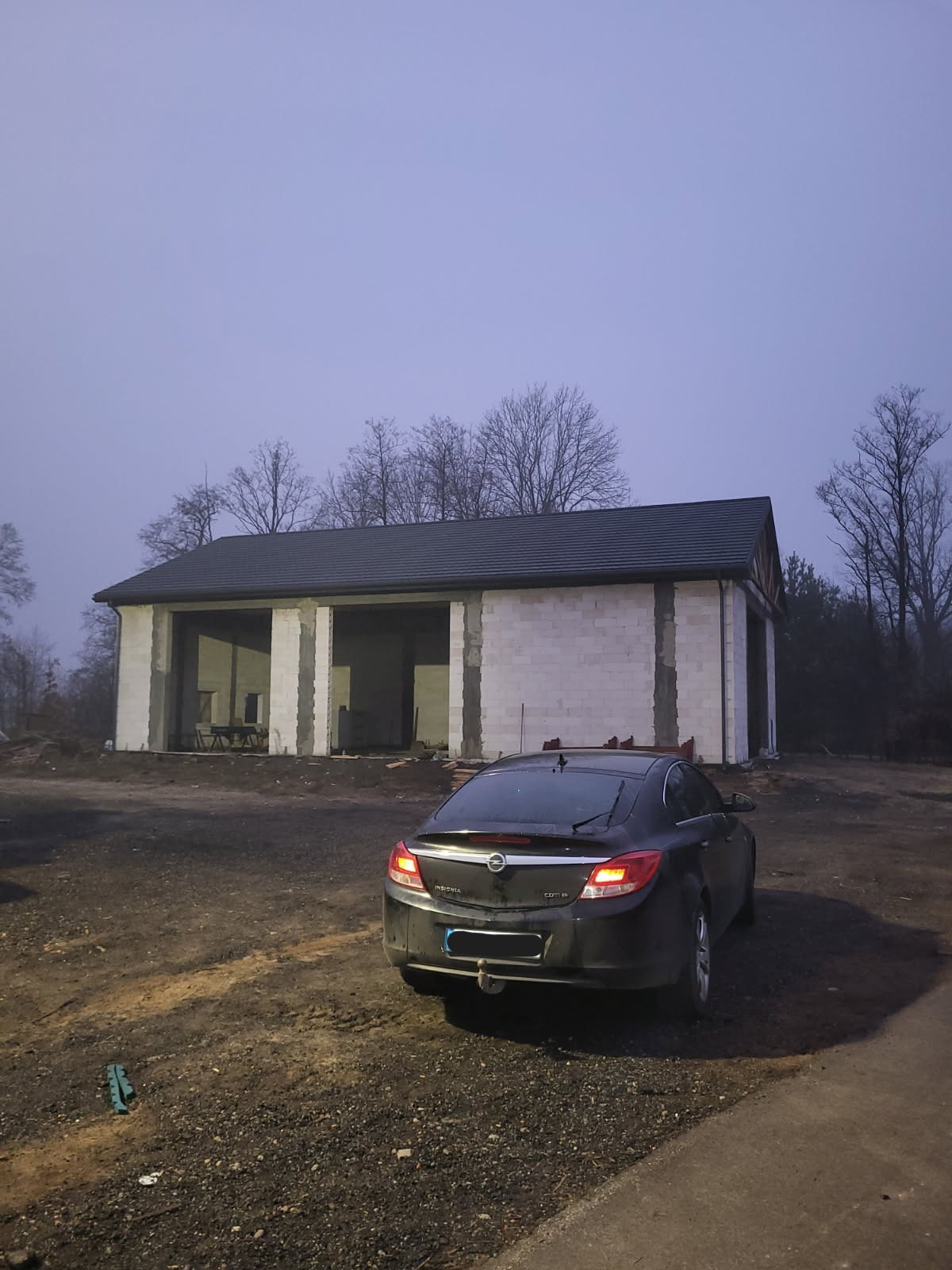 Surowy budynek z białego bloczka, z ciemnym dachem, w tle drzewa bez liści, na pierwszym planie zaparkowany czarny samochód osobowy, pochmurny dzień.