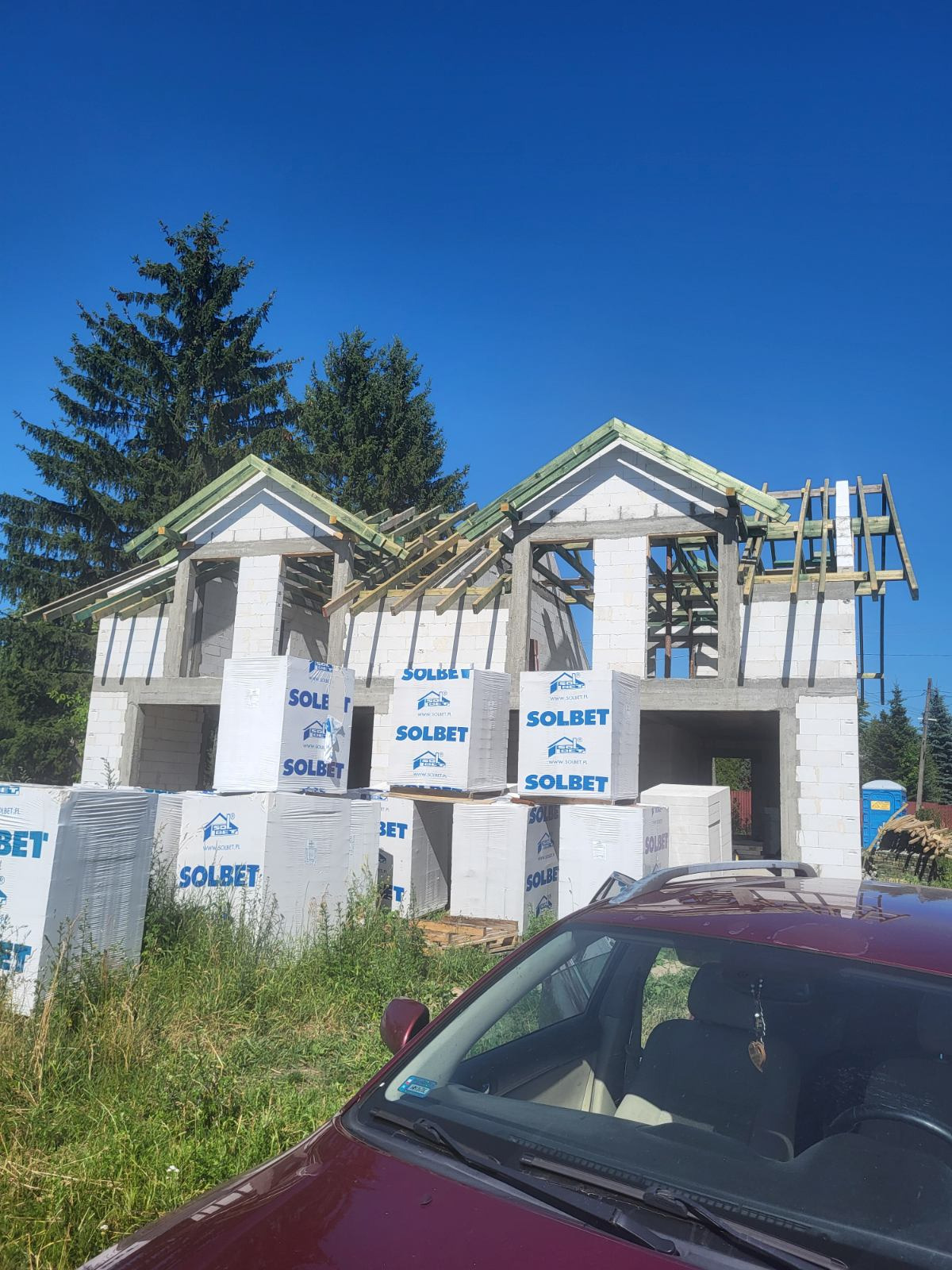 Budowa domu z białych bloczków Solbet, widoczne drewniane elementy konstrukcji dachu. Na pierwszym planie zapakowane bloczki i fragment samochodu.