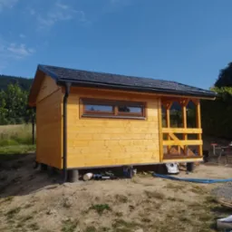 Żółty, drewniany domek narzędziowy z czarną, spadzistą dachówką, z małym oknem i zadaszonym wejściem, na tle błękitnego nieba i zieleni. Widoczne narzędzia.